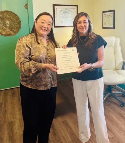 Lopez Law Office, P.C. Selected for Mexican Consulate Attorney Referral Network Two women standing indoors, smiling and holding a certificate together. Framed diplomas are visible on the wall behind them, and a dog is lying in a bed on the floor.
